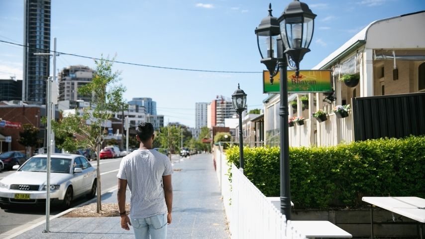 A man walks along a sunny city sidewalk past a white car, modern buildings, and a café terrace with ornate black street lamps, evoking a calm urban afternoon.