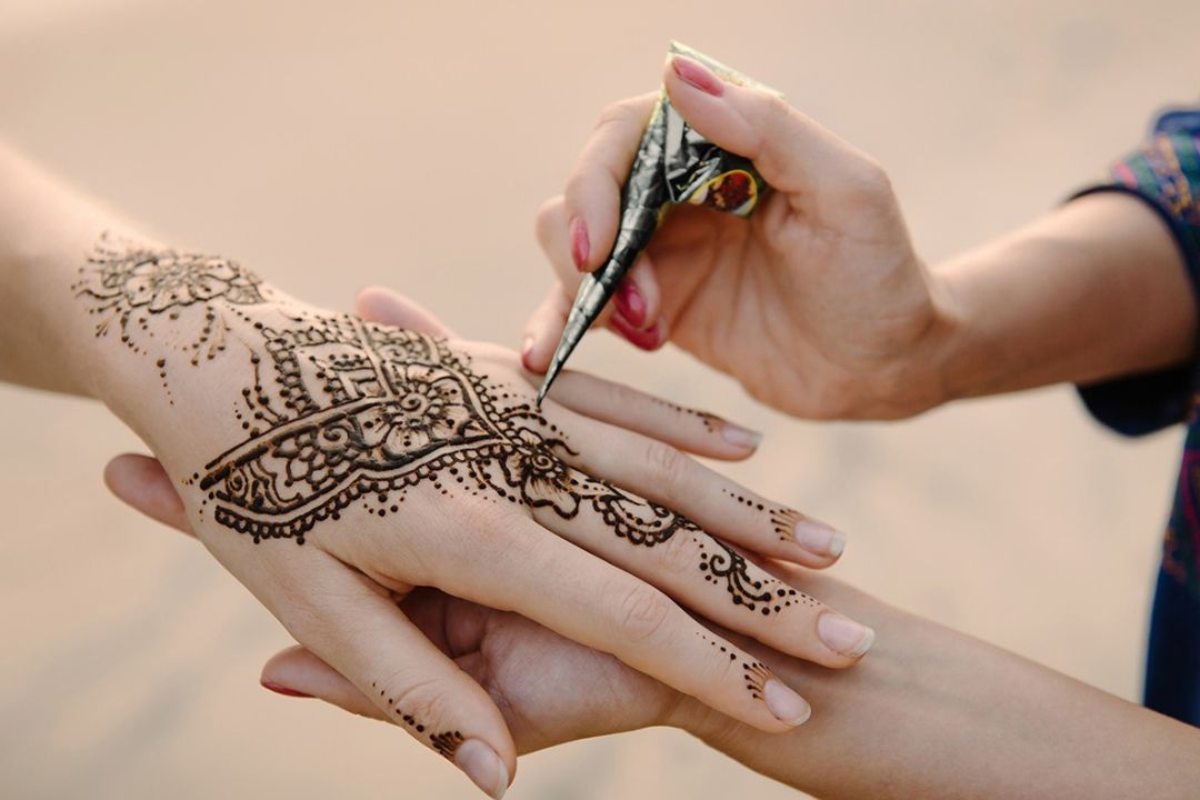 A henna artist applies intricate dark floral and lace patterns to a woman’s hand, creating an intimate, careful moment of traditional body art.