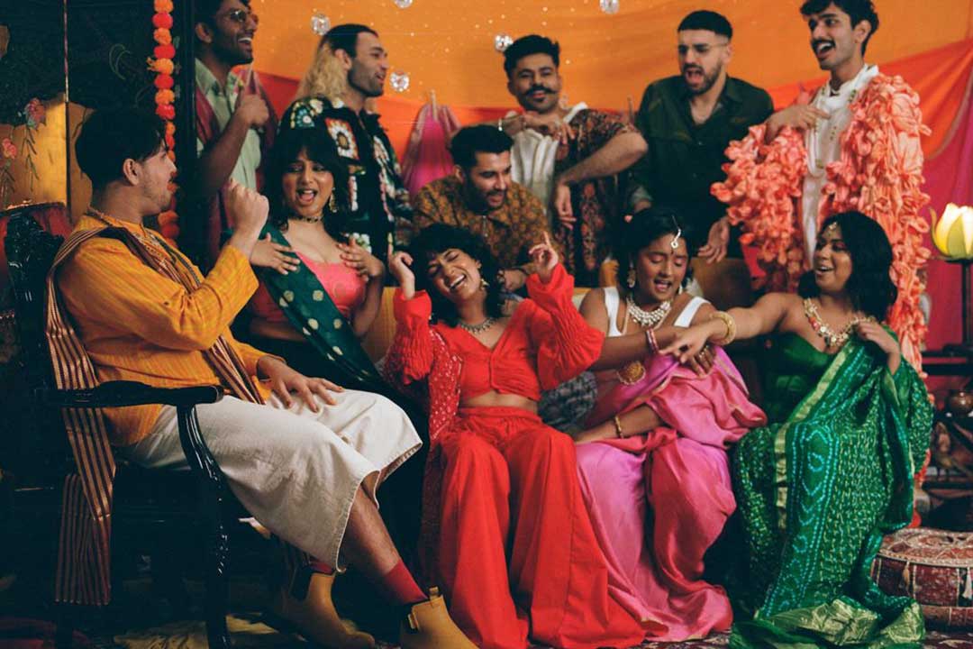 Friends in colorful traditional outfits laugh, sing, and dance at a festive gathering, framed by orange drapes, flower garlands, and warm decor.