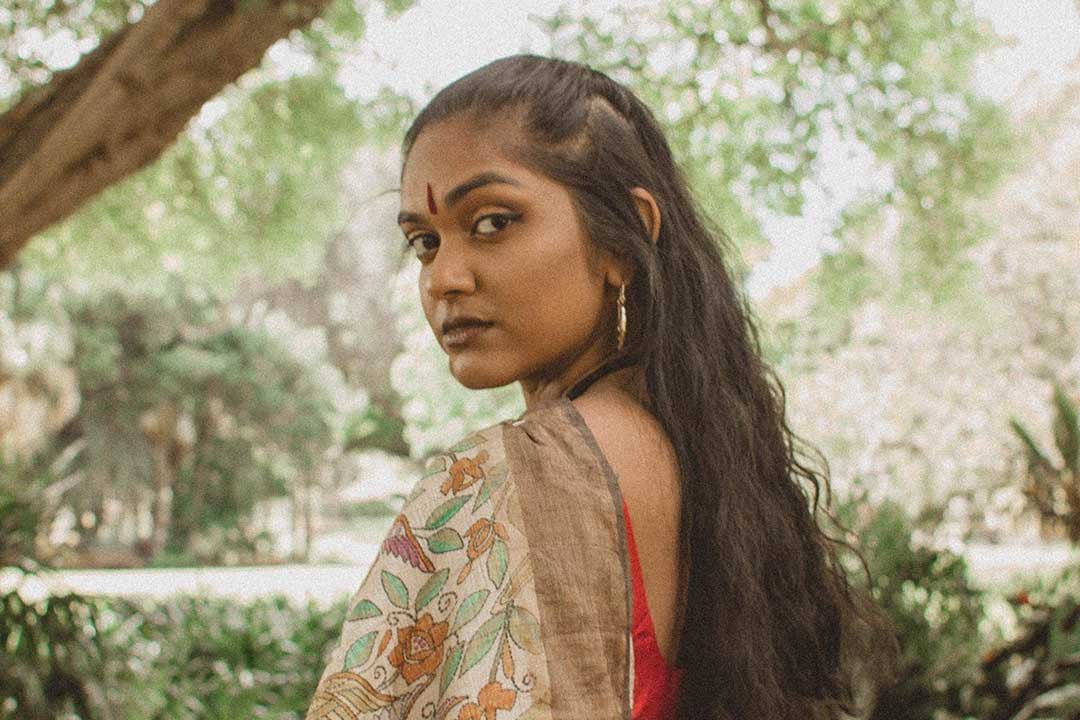 A woman in a floral sari looks calmly over her shoulder beneath leafy trees, her long dark hair falling down her back in soft outdoor light.