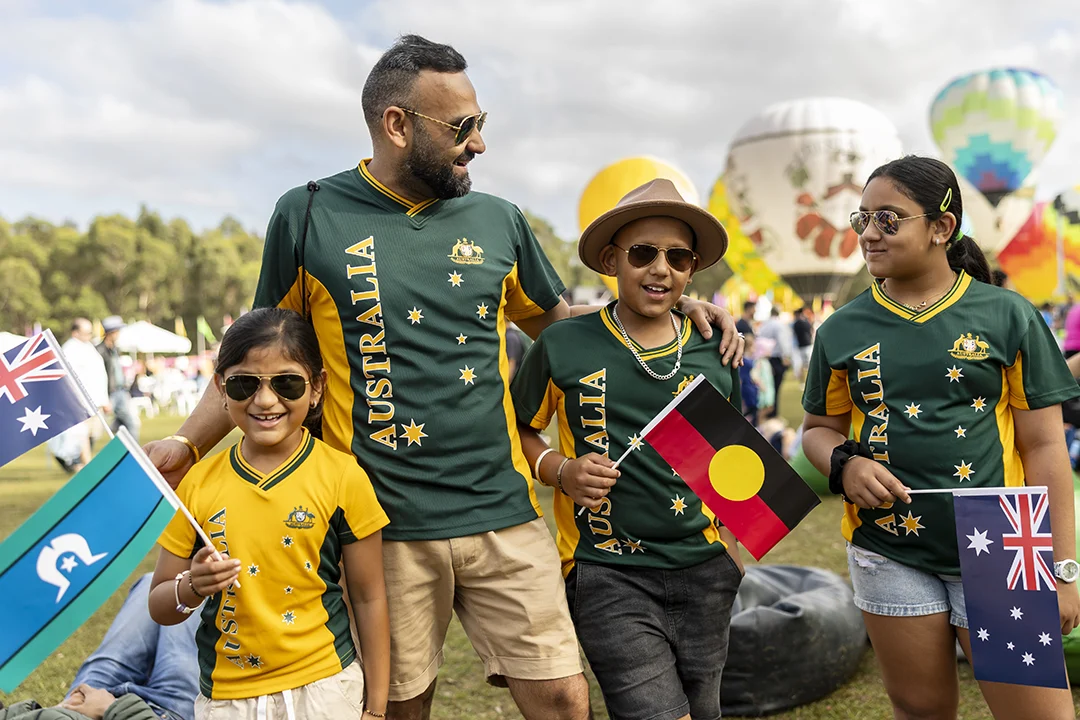 A smiling family in green and yellow Australia jerseys poses at an outdoor festival, holding Australian and Aboriginal flags beneath colorful balloons.
