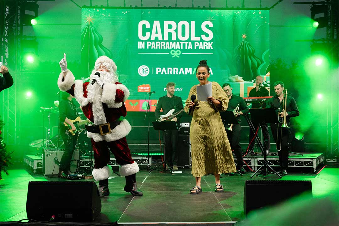 Santa dances beside a smiling singer in a gold dress on a green-lit stage with a live band, beneath a “Carols in Parramatta Park” sign.