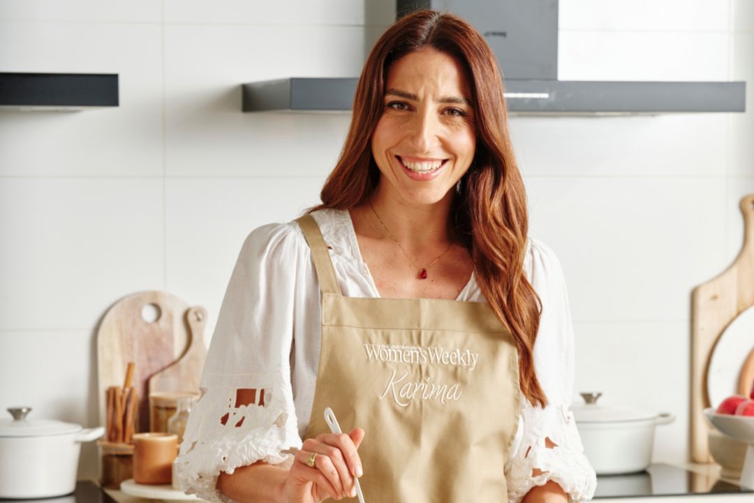 A smiling red-haired woman in a beige apron holds a wooden spoon in a bright white kitchen, with cookware and wooden boards behind her.