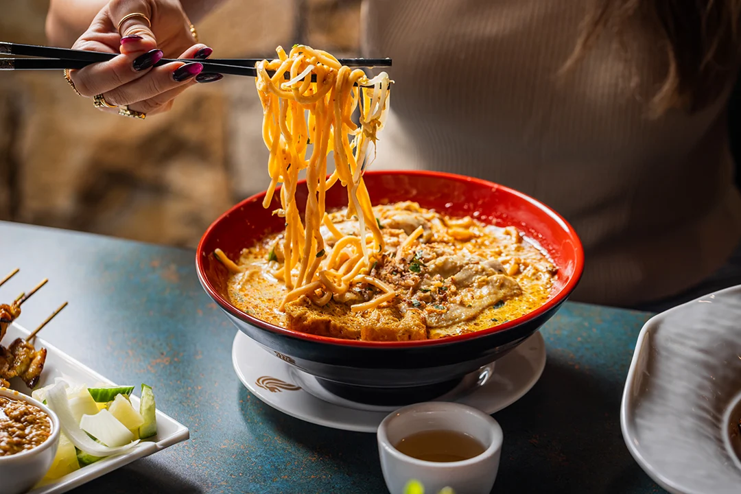 A hand lifts glossy noodles with chopsticks from a red bowl of spicy broth at a restaurant table, evoking a warm, appetizing meal.