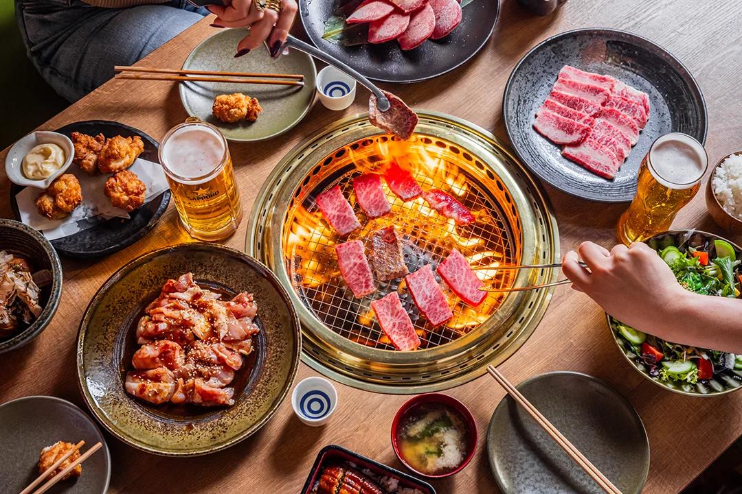 Overhead viewJapanese barbecue, with diners grilling marbled beef over glowing charcoal, surrounded by raw meat, marinated pork, beer, and side dishes.