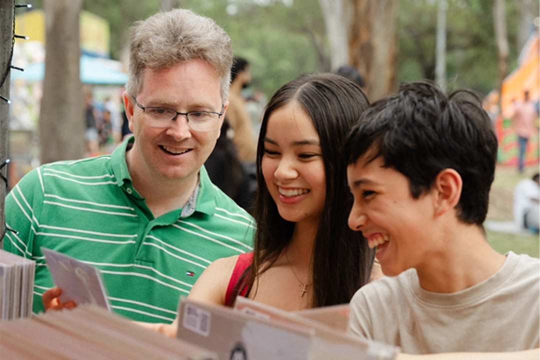 A middle-aged man and two young adults smile as they study a brochure outdoors at a lively fair, sharing a warm, cheerful moment.