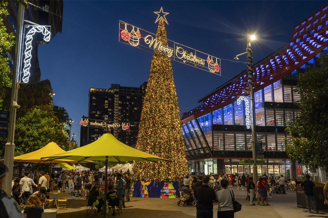 Crowds gather at a Christmas market in Parramatta Square at night, with a towering Christmas tree covered in golden lights and ornaments at the centre.