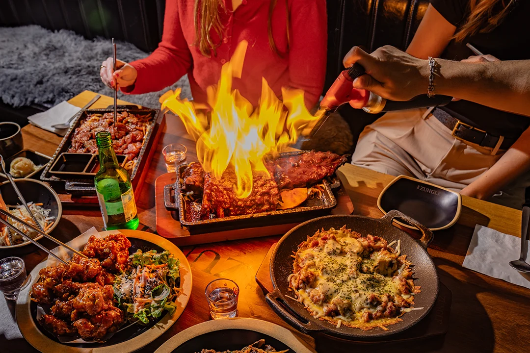 Diners gather around a lively barbecue spread as flames rise from a platter of grilled meat, with side dishes, drinks, and warm restaurant lighting.