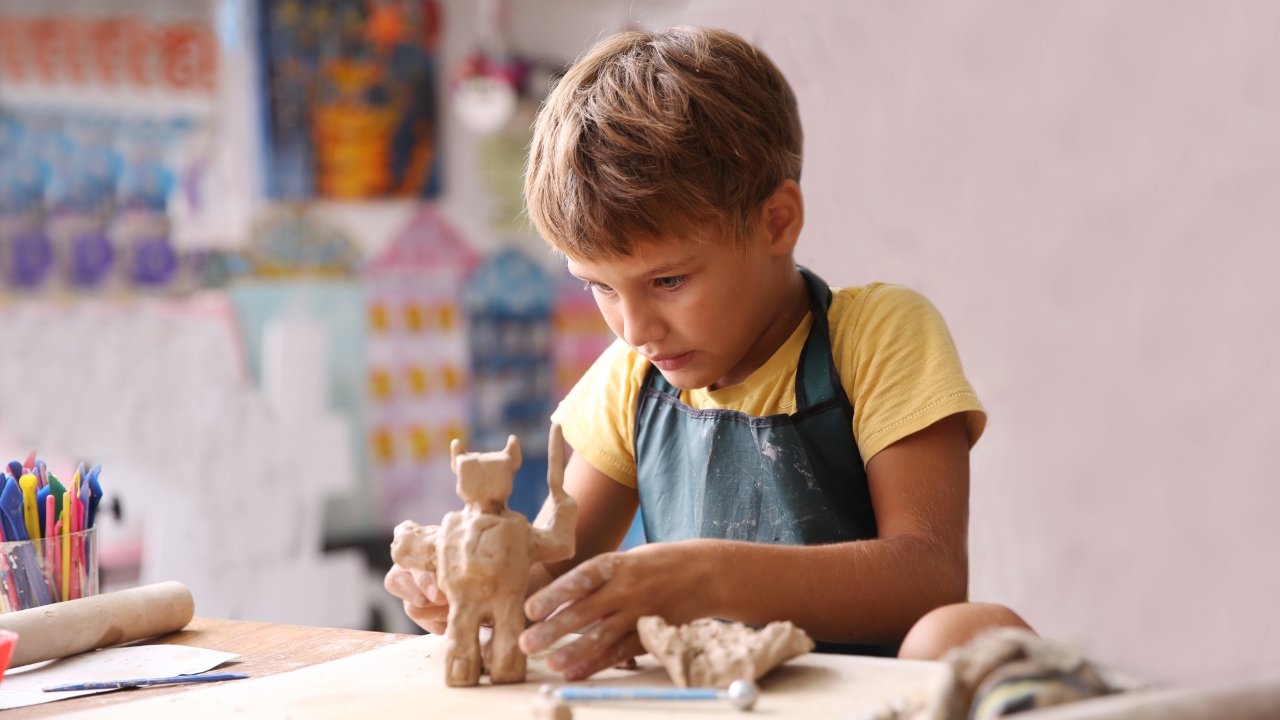 A focused child in a yellow shirt and apron sculpts clay at a table, surrounded by colourful art supplies, in a vibrant, creative studio setting.