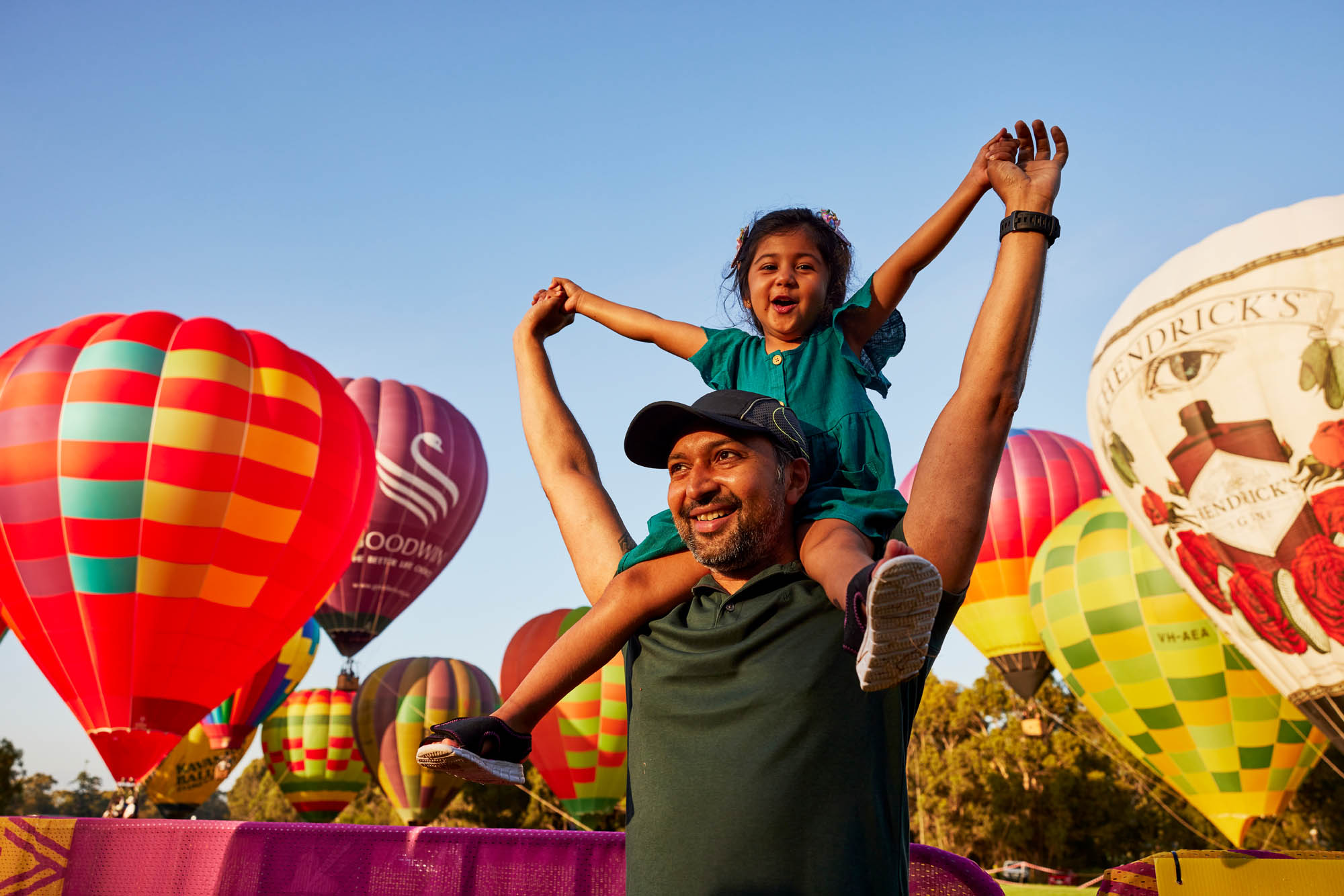 Child on man shoulders with hot air balloons in background.