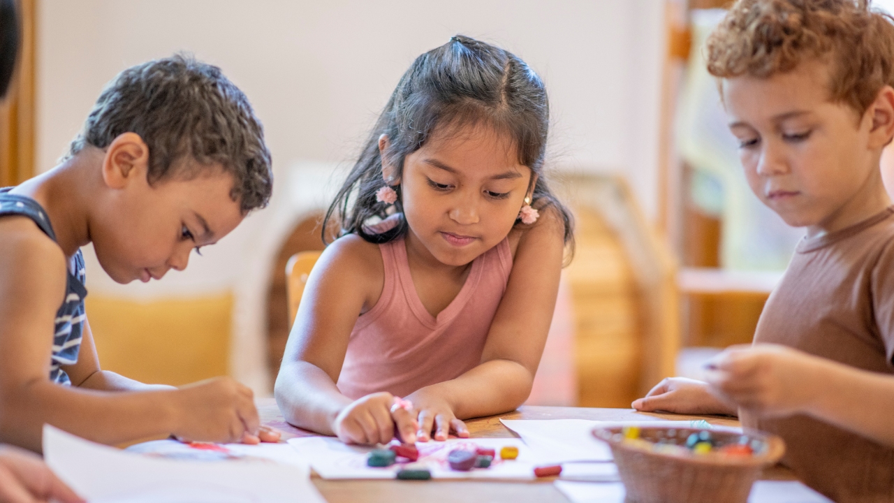 Three children are sitting around a table, drawing with crayons on paper. They appear focused and engaged, sharing colors from a basket.