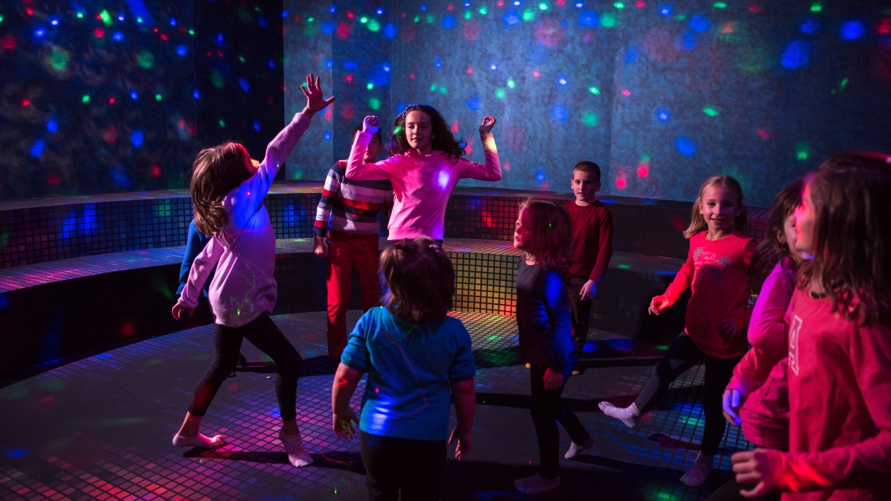 Children joyfully dance under colorful disco lights in a dark room with a curved bench. The scene is lively, capturing a playful and energetic atmosphere.
