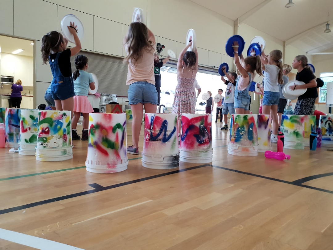 Children are inside a gym, each holding colorful buckets and plates, engaged in a lively drum circle. The mood is playful and energetic.
