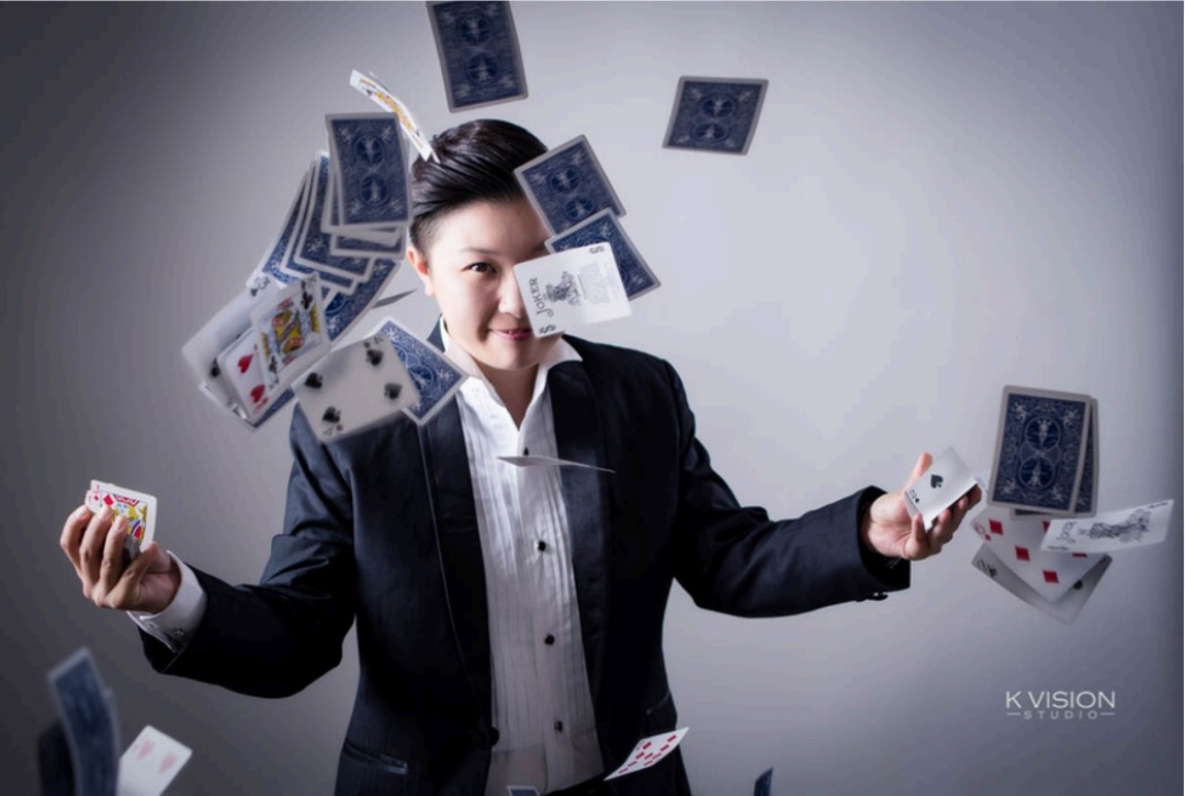 A person in a suit joyfully juggles playing cards in mid-air against a neutral background. The scene conveys a sense of magic and skillful performance.