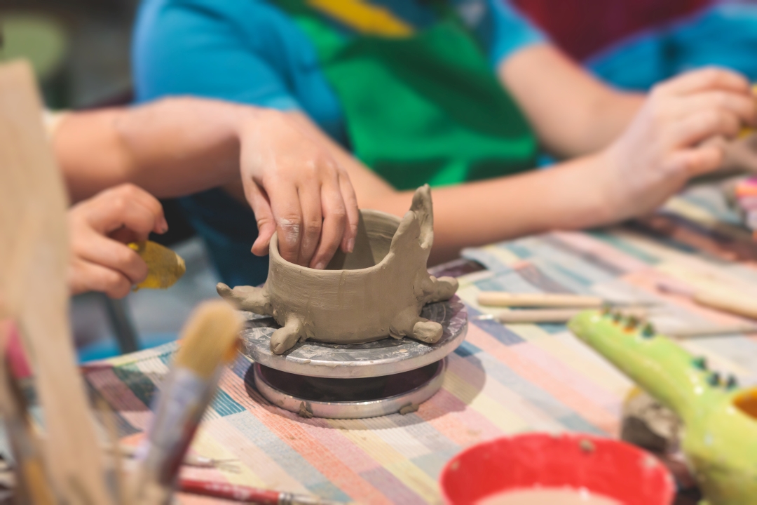Children shaping clay into a bowl on a colorful tablecloth, surrounded by brushes and tools. The scene conveys creativity and focus in a pottery class.