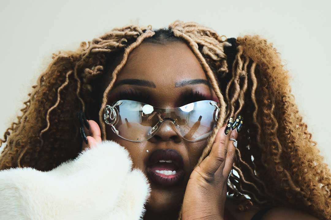 A woman with blonde braids and bold makeup, wearing sunglasses and a white fur coat.