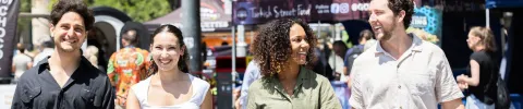 Four people walk side by side in a lively outdoor street food market. They are smiling and enjoying the sunny day, creating a cheerful and friendly atmosphere.
