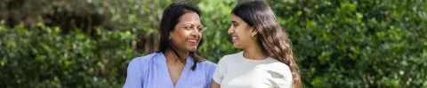 A woman and a young girl smile warmly at each other while walking in a lush green garden, conveying a sense of joy and connection.