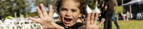 A joyful child with curly hair smiles widely and stretches out hands toward the camera at an outdoor event. Background shows chairs and trees under a sunny sky.