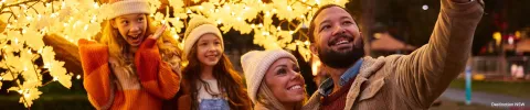 A joyful family of four, dressed warmly in coats and beanies, smile under glowing tree lights in a nighttime setting, conveying warmth and happiness.