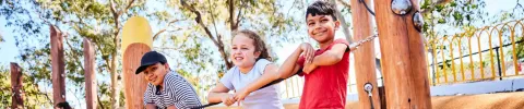 Three children balance on a wooden beam at a playground, smiling and holding onto ropes. Sunlight filters through trees, creating a cheerful mood.
