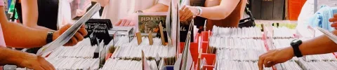 Hands browse through rows of vinyl records in colorful bins at a busy market. The scene conveys a nostalgic and lively atmosphere.