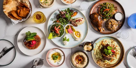 Overhead spread of colorful Middle Eastern dishes, grilled meats, salads, hummus, flatbread, sauces, and drinks on a marble table.