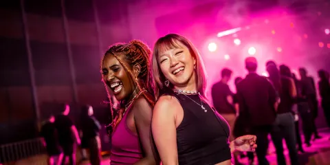 Two joyful women dance back-to-back at a lively nightclub, bathed in vivid pink and purple lights, with a silhouetted crowd behind them.