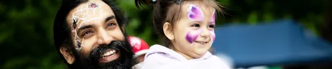 A smiling bearded man with face paint holds a young child with purple and pink cheek designs outdoors; their joyful expressions create a playful festival mood.