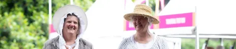 Two people wearing historical-style clothing and wide-brimmed hats stand at an outdoor community event.