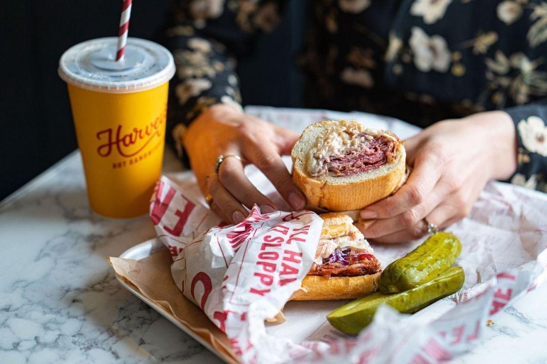 Person sat at a table holding a Harvey's hot sandwiches with a drink