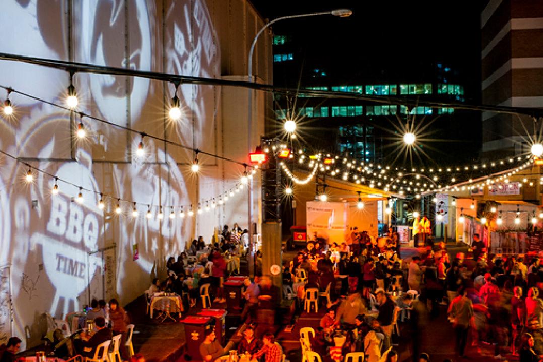 A laneway in Parramatta is activated during the evening with lights, food trucks, live music and murals projected against the wall. 