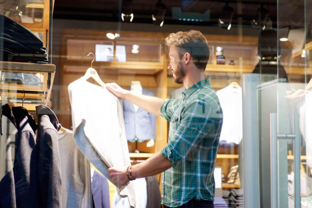 Image of a male shopper in a retail clothing store. He is dressed casually holding up a dress shirt to inspect it. The store atmosphere is warm and inviting like a shop patrons would enjoy spending time in. 