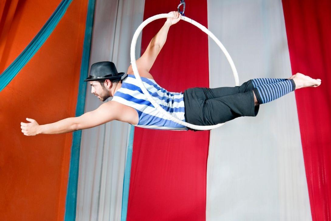 man practicing circus skills, suspended in giant hoop in the air with circus tent behind him