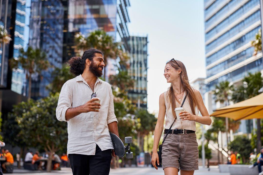 two students walking and talking to each other in parramatta square, both people are holding a coffee and one of them is holding a skateboard under their arm
