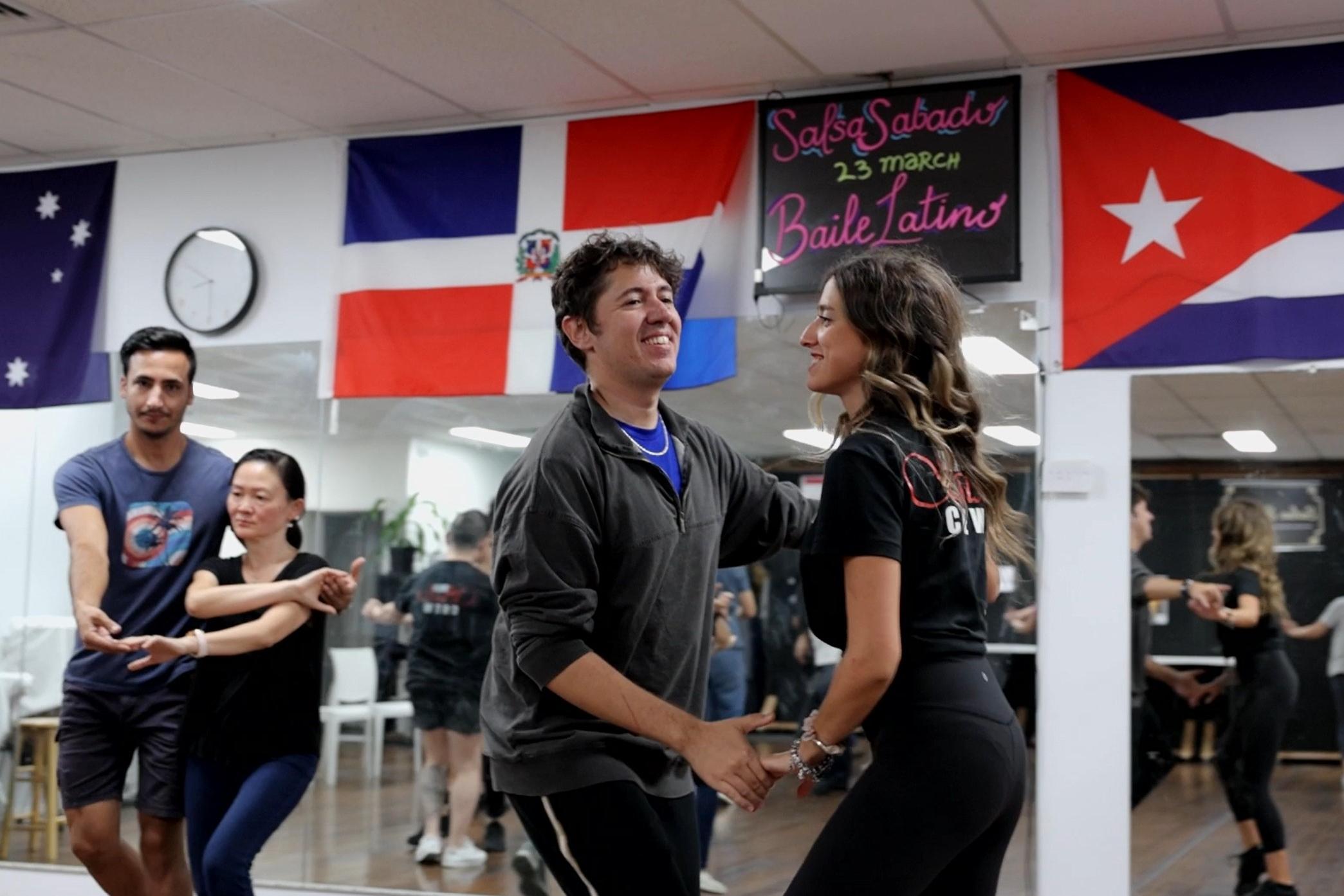 Couples dance joyfully in a lively class, surrounded by international flags, including France and Cuba, creating a vibrant, multicultural atmosphere.