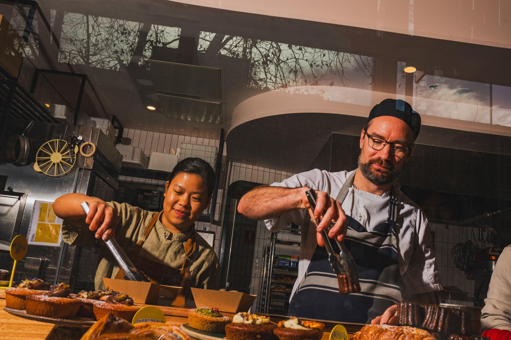 Two people holding tongs, serving food at a cafe counter