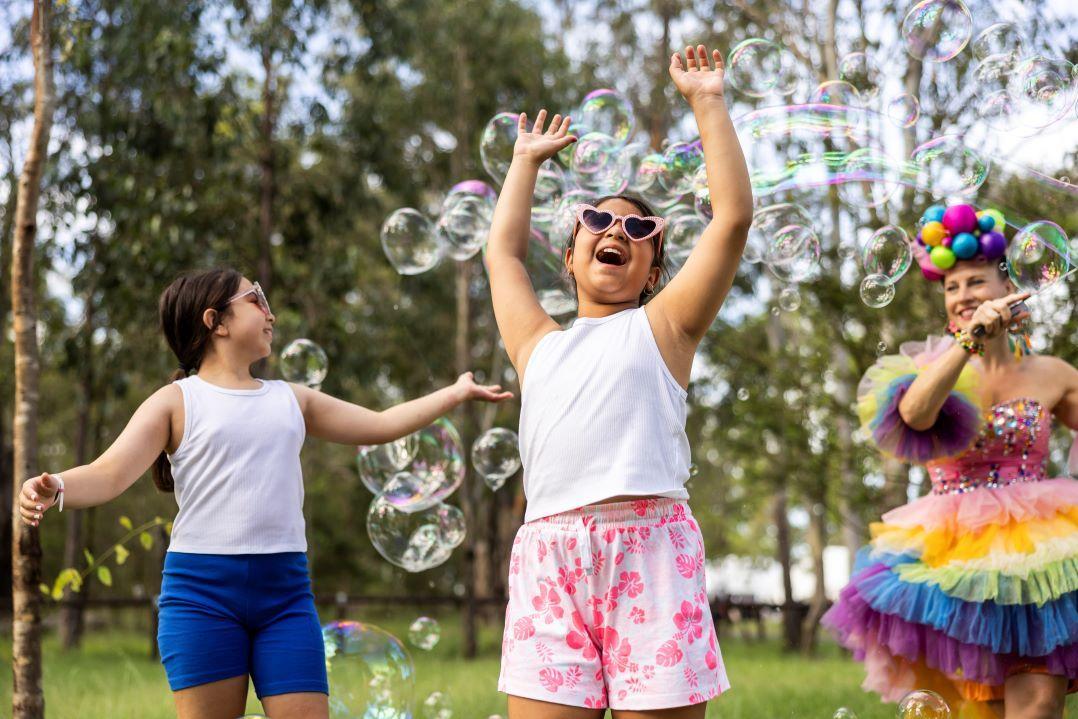 Three people outdoors joyfully dance in colorful clothes. One wears sunglasses and pink shorts, another a rainbow tutu. They are surrounded by trees.