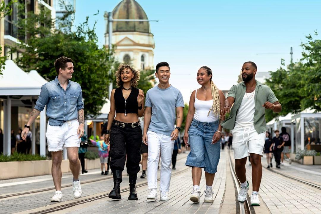 A diverse group of five young adults joyfully walk down a sunny, tree-lined street, holding hands. Tents and a domed building are visible in the background.