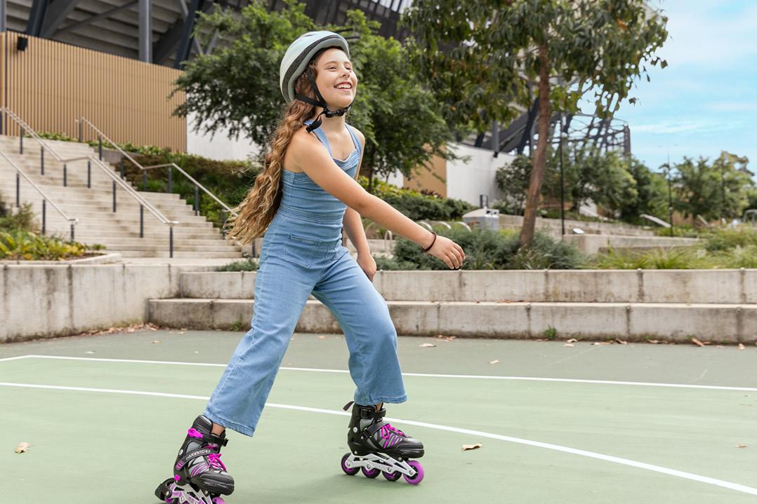 A smiling girl in a helmet and overalls rollerblades on an outdoor court, surrounded by trees and greenery, conveying a sense of joy and freedom.