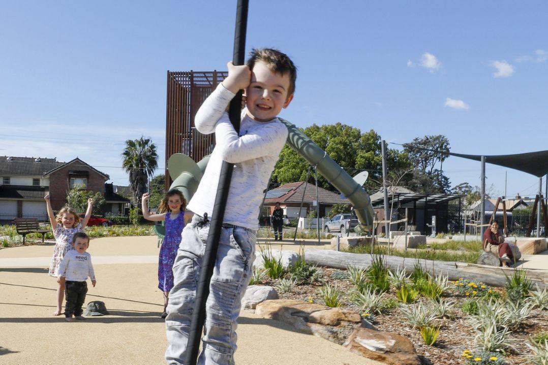 A child in a gray shirt joyfully plays on a pole in a sunny park. In the background, another child walks, and green trees dot the landscaped area.