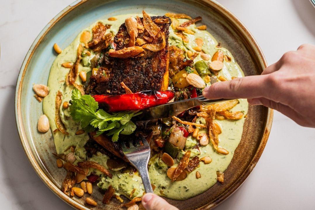 A vibrant salad on a blue plate with mixed greens, sliced avocado, red peppers, and crispy onions. Two hands are reaching to serve the salad, suggesting a communal meal.