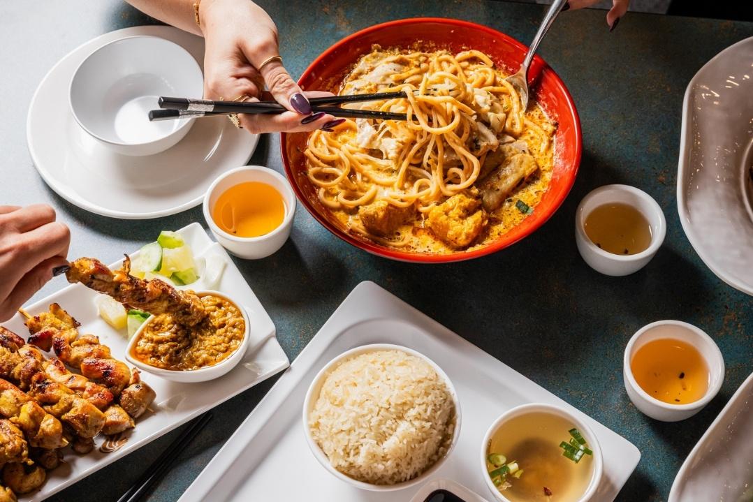 A hand serves pasta from a red bowl, surrounded by plates of fried chicken, coleslaw, and sauces. Warm tones suggest a cozy dining atmosphere.