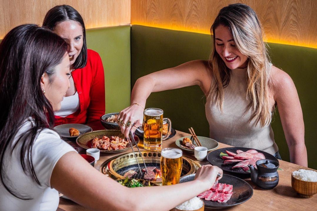 Three women enjoying a meal at a restaurant, each with local cuisine. They are smiling, conveying a sense of friendship and enjoyment.