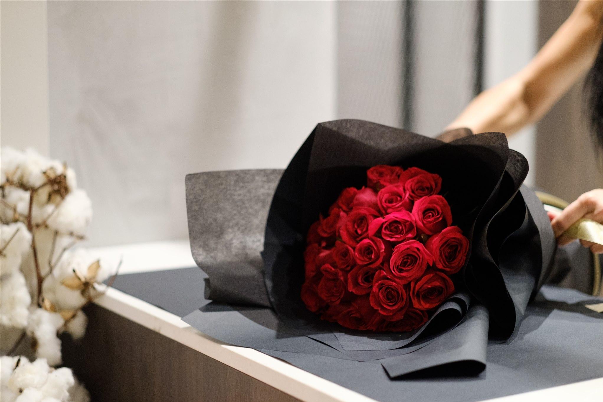 A bouquet of vibrant red roses wrapped in black paper is being held by a person. Cotton buds in the background add a soft, elegant contrast.