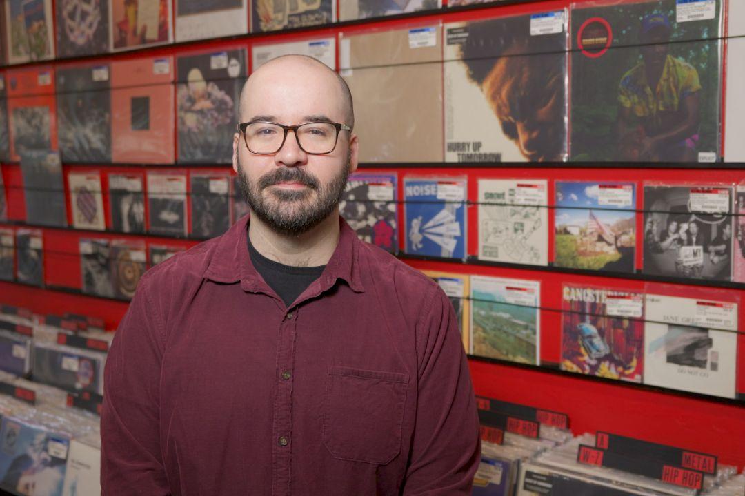 Bald man in a maroon shirt and glasses sitting in front of a walk of records