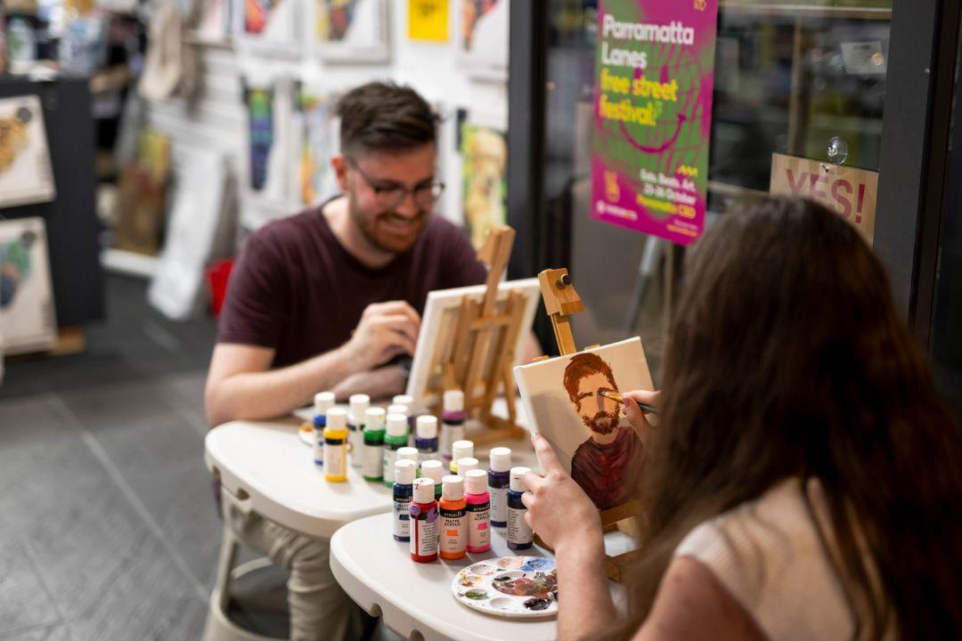 Man and woman sitting at small table with art canva infront of them painting each other
