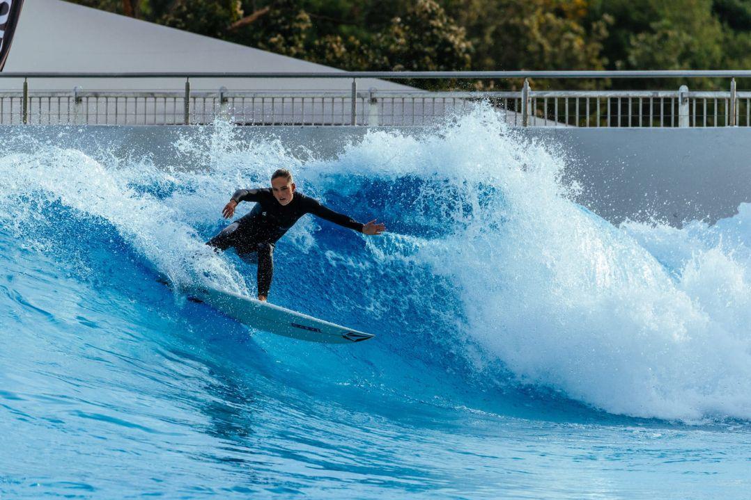 A surfer in a black wetsuit rides a dynamic wave in an artificial wave pool. The scene conveys excitement and skill, with foamy water splashing around.