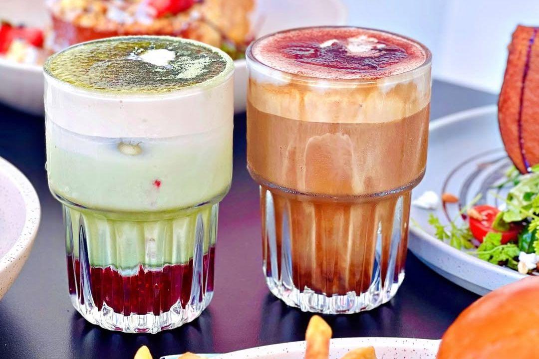 2 glass cups. One with green matcha with raspberries on the bottom, and second cup with coffee and raspberry on top