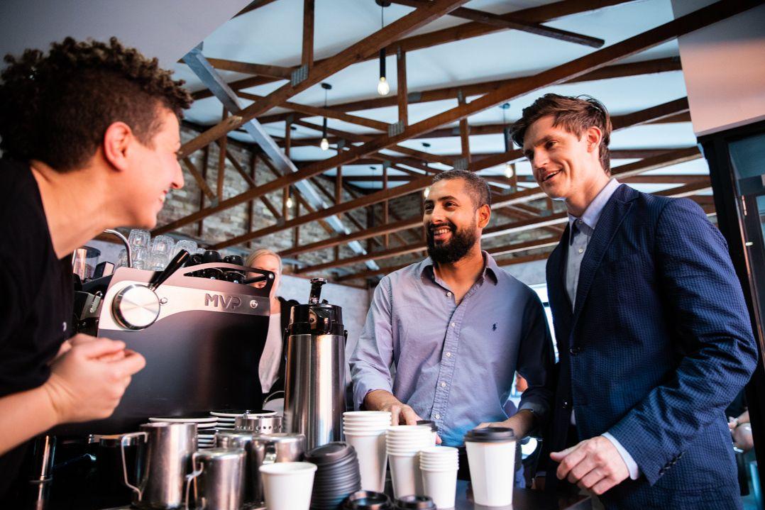 Two business man in suits smiling at lady standing behind coffee machine making drinks